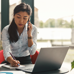 Woman with dark long hair in a ponytail speaking on a mobile phone while writing on a notepad in front of a laptop. 