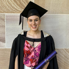Bachelor of Bachelor of Arts in the field of Psychology Graduate Casey Gilbert in their graduation robes and hat against a sandstone wall