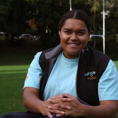 A young woman sitting on grass in front of a park with AFL posts in the background
