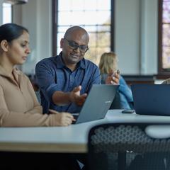 An older male teaching a male and female student at a desk on their laptops