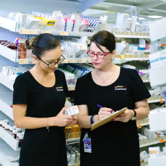 Two pharmacists in black uniforms review a medication box and make notes on a clipboard, standing in front of shelves filled with various pharmacy products.