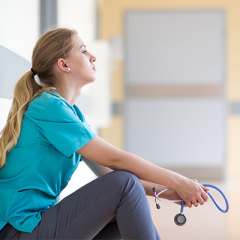 Female nurse sitting on ground against wall. She has long blonde hair in a ponytail and is wearing a blue shirt and grey pants. 