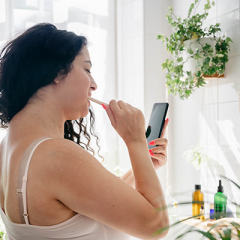 Woman with dark hair brushing teeth while watching phone 