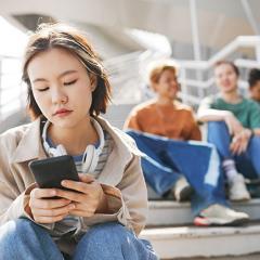 A young woman sitting on steps, engrossed in her phone with 3 people chatting behind her.
